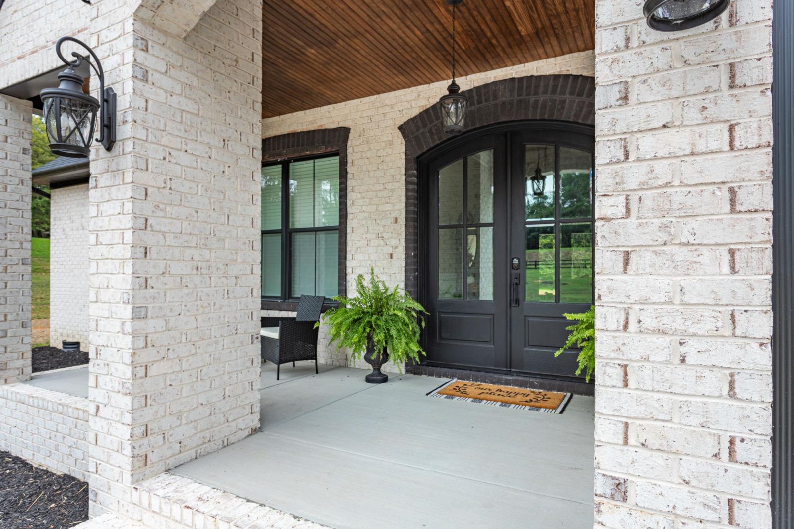 A double sided door on a bricked porch, with lantern style lights on each side of the door as well as shrubs below them on each side of the doormat