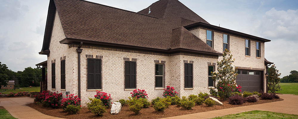 A white brick home with roses in the surrounding flowerbeds, with a mocha roof