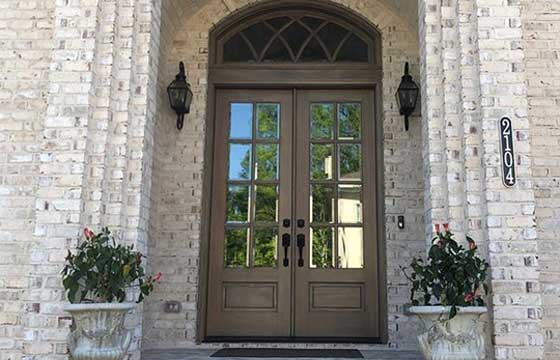 A double sided door on a bricked porch, with lantern style lights on each side of the door as well as shrubs below them on each side of the doormat