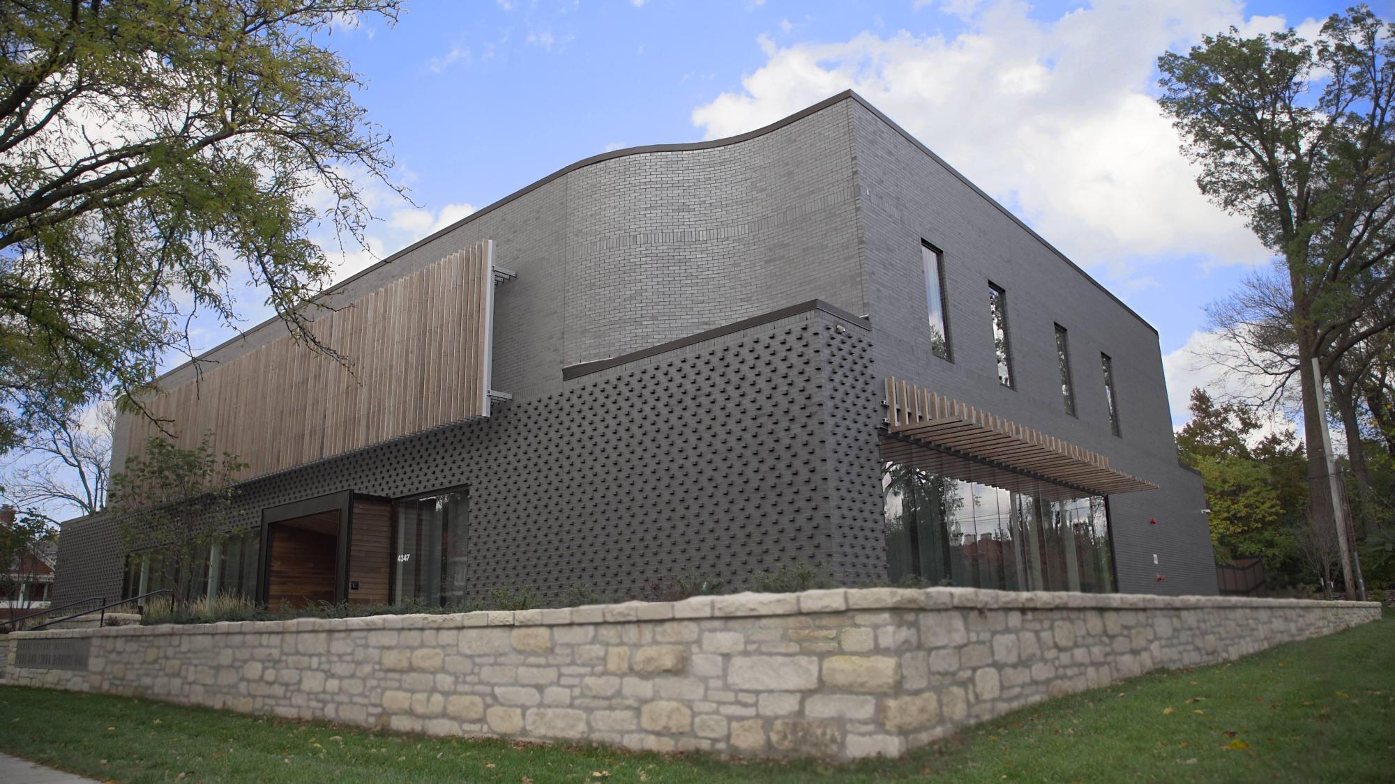 Image of Paul and Linda DeBruce Hall; gray bricked and surrounded by a stone wall, trees, and lush grass