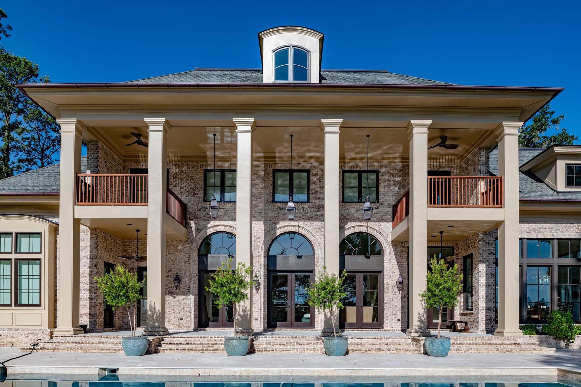 Image of the front of a custom home, pine trees shadow a shingled roof held aloft by tall pillars shrouded by potted trees